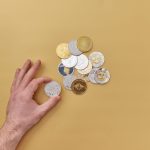 A hand picks cryptocurrencies from a mix of gold and silver coins on a vibrant yellow background. — Bitcoin Price and Its Imp