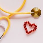 Top view of yellow stethoscope made of plastic and metal material near small handmade heart of spiral hair tie on pale pink b