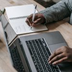 Crop unrecognizable African American female in formal wear writing notes in notepad while working on laptop — India Now - The