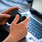 Person holds a gaming controller while interacting with a laptop, emphasizing modern gaming technology. — Information Media I