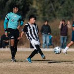 A youth soccer player strikes the ball during a match with a referee nearby, expressing focus and teamwork. — La Grada Online