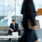 Businessman sitting in modern office lounge using a laptop while colleague walks past, creating dynamic blur. — Looking Into