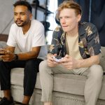 Two friends sitting on a sofa, playing video games together at home. — Newspapers - The Crucial Part of Journalism