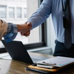 Crop unrecognizable coworkers in formal wear standing at table with laptop and documents while greeting each other before mee